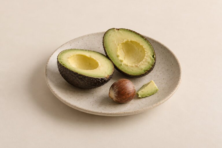 A ripe avocado, a small piece of flesh and its pit sit apart on a plain ceramic plate.