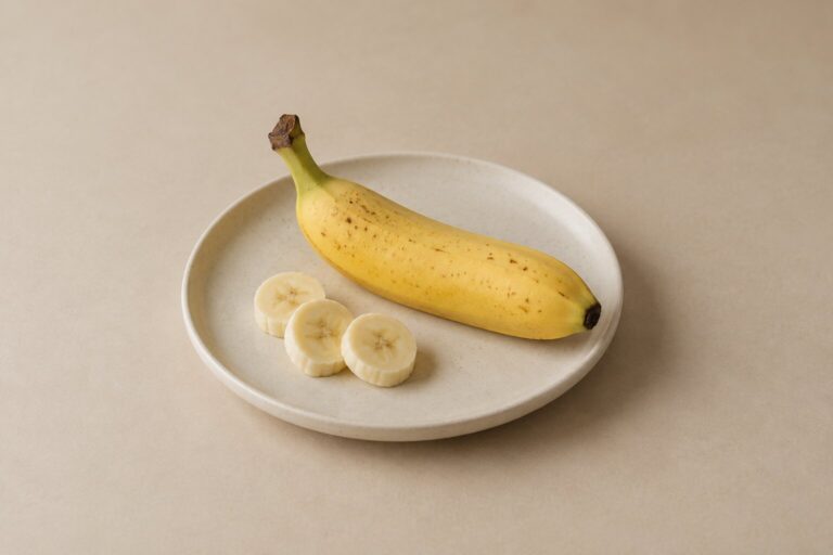 A ripe banana and three thin slices rest on an ivory plate against an oat backdrop.