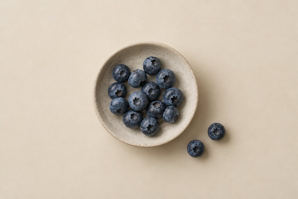 Fresh blueberries sit in a shallow stoneware dish with two berries beside it on a warm bone background.