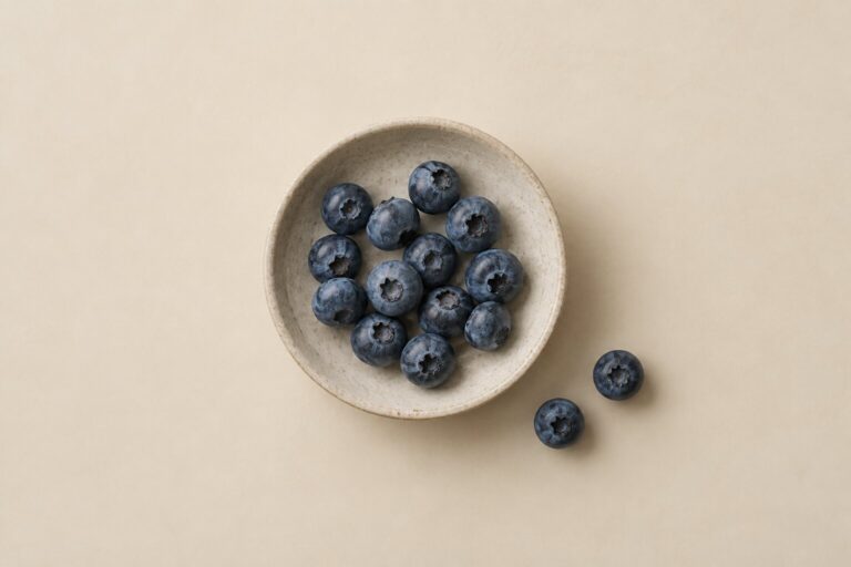 Fresh blueberries sit in a shallow stoneware dish with two berries beside it on a warm bone background.