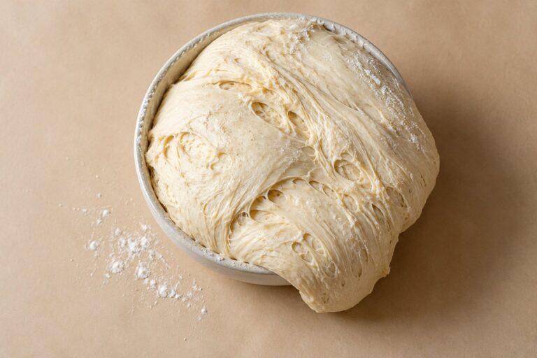 A ball of raw bread dough spills over the edge of a ceramic bowl on a beige backdrop.