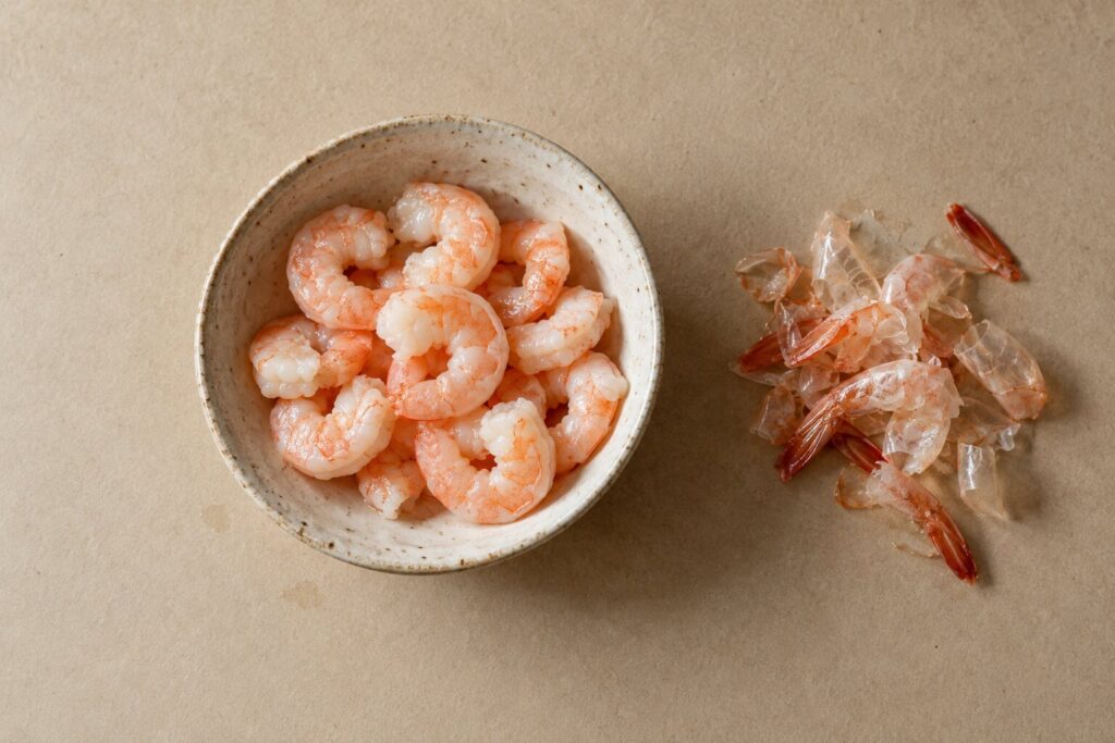 Bowl of cooked peeled shrimp pieces beside a small pile of discarded shrimp shells and tails